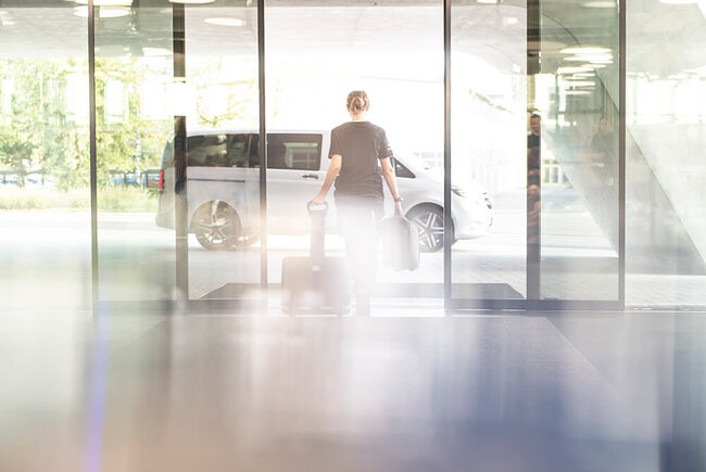 A young woman with a suitcase on wheels is leaving a modern building.