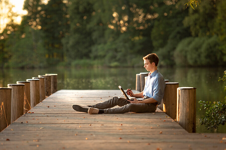 Ein junger Mann sitzt mit einem Notebook auf einem Bootssteg.