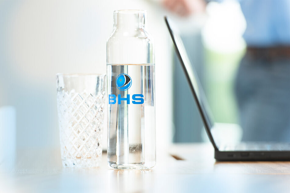 A fancy water bottle with the BHS-Corrugated logo and a glass.