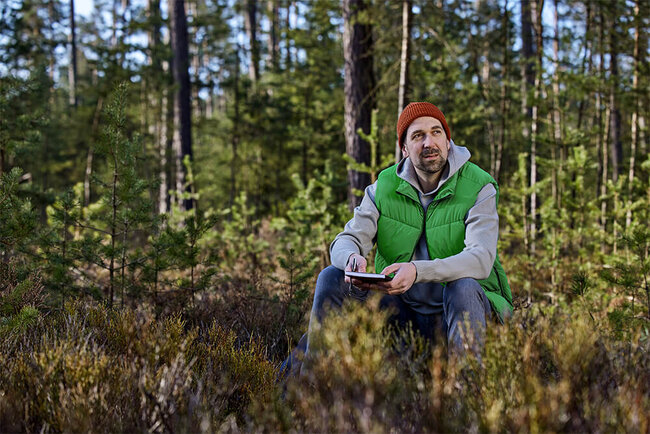 Wolfgang Seegerer sitzt in einem Waldstück. Er hält ein Tablet in der Hand.
