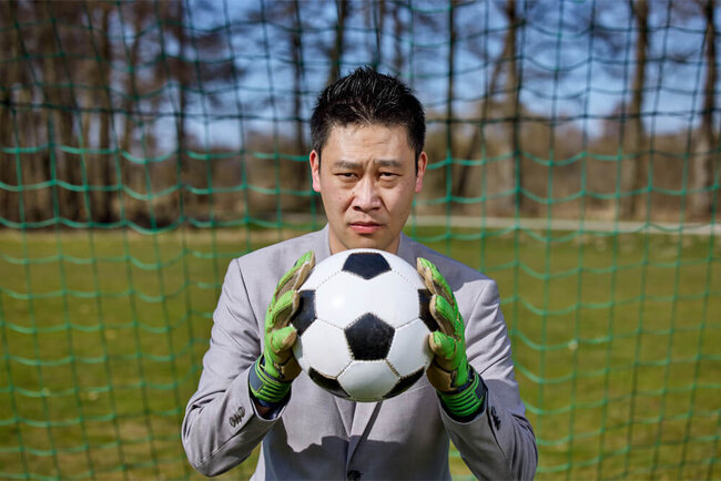 Kui Hou steht in der Mitte eines Fußballtors. Er hält einen Fußball und blickt direkt in den Kamera.