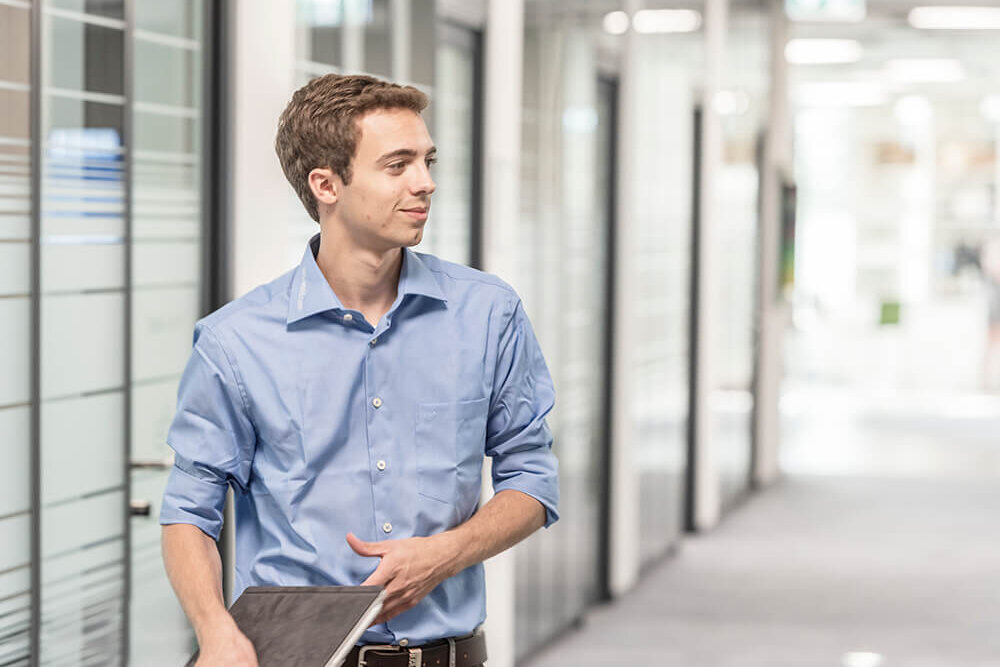 Ein junger Mann geht mit einem Notebook unterm Arm durch ein Firmengebäude.