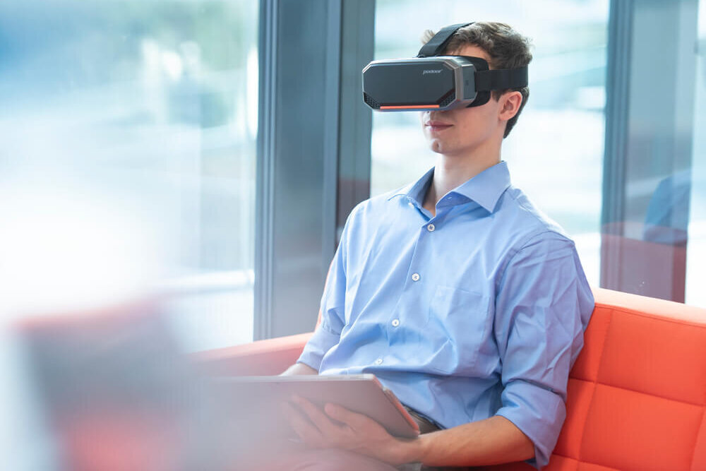 A youg man is holding a tablet. He is wearing a virtual reality headset.