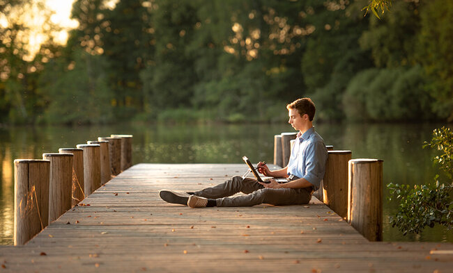 Ein junger Mann sitzt mit seinem Notebook auf einem Holzsteg an einem See.